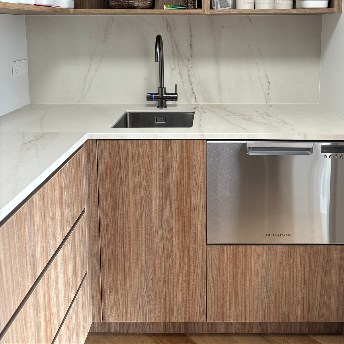 A modern kitchenette corner with light wood grain cabinets, a white marble-look countertop, a built-in stainless steel dishwasher, and open upper shelves holding kitchen items.