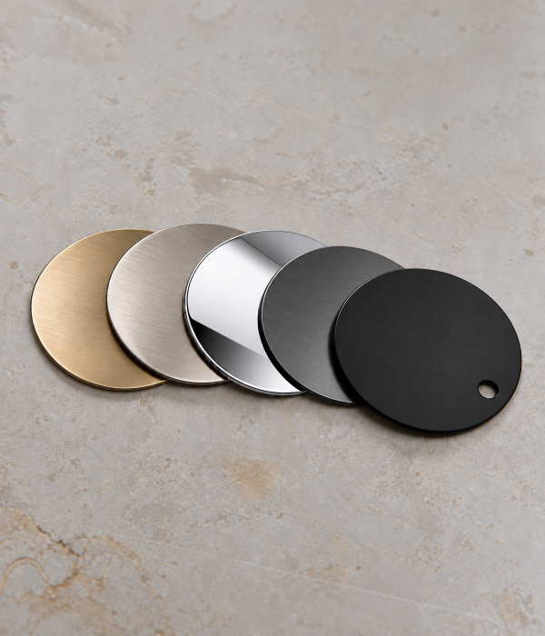 Five round metal tags or plates in different finishes—brass, brushed silver, polished silver, matte gray, and matte black—are arranged in a row on a light beige stone surface.