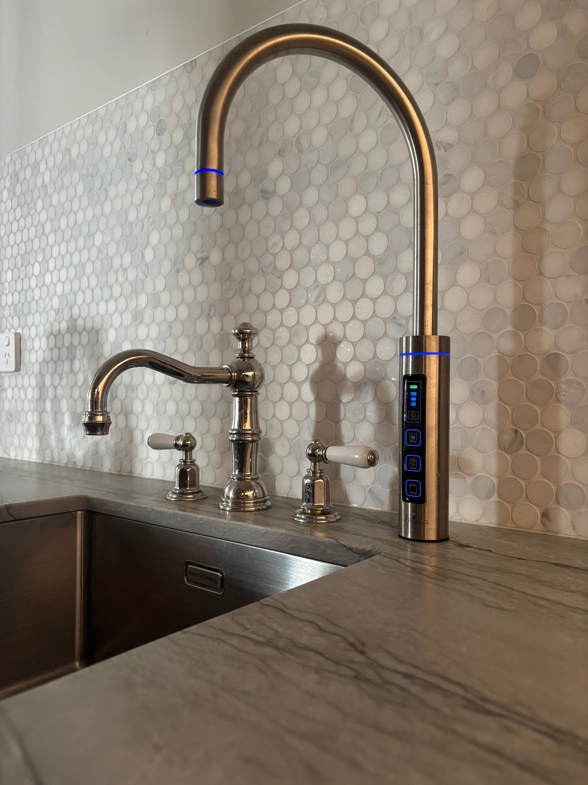 A modern wall-mounted kitchen faucet set with a curved gooseneck sprayer, a separate curved spout, and a digital control panel, installed against a white hexagonal tile backsplash and a gray stone countertop.